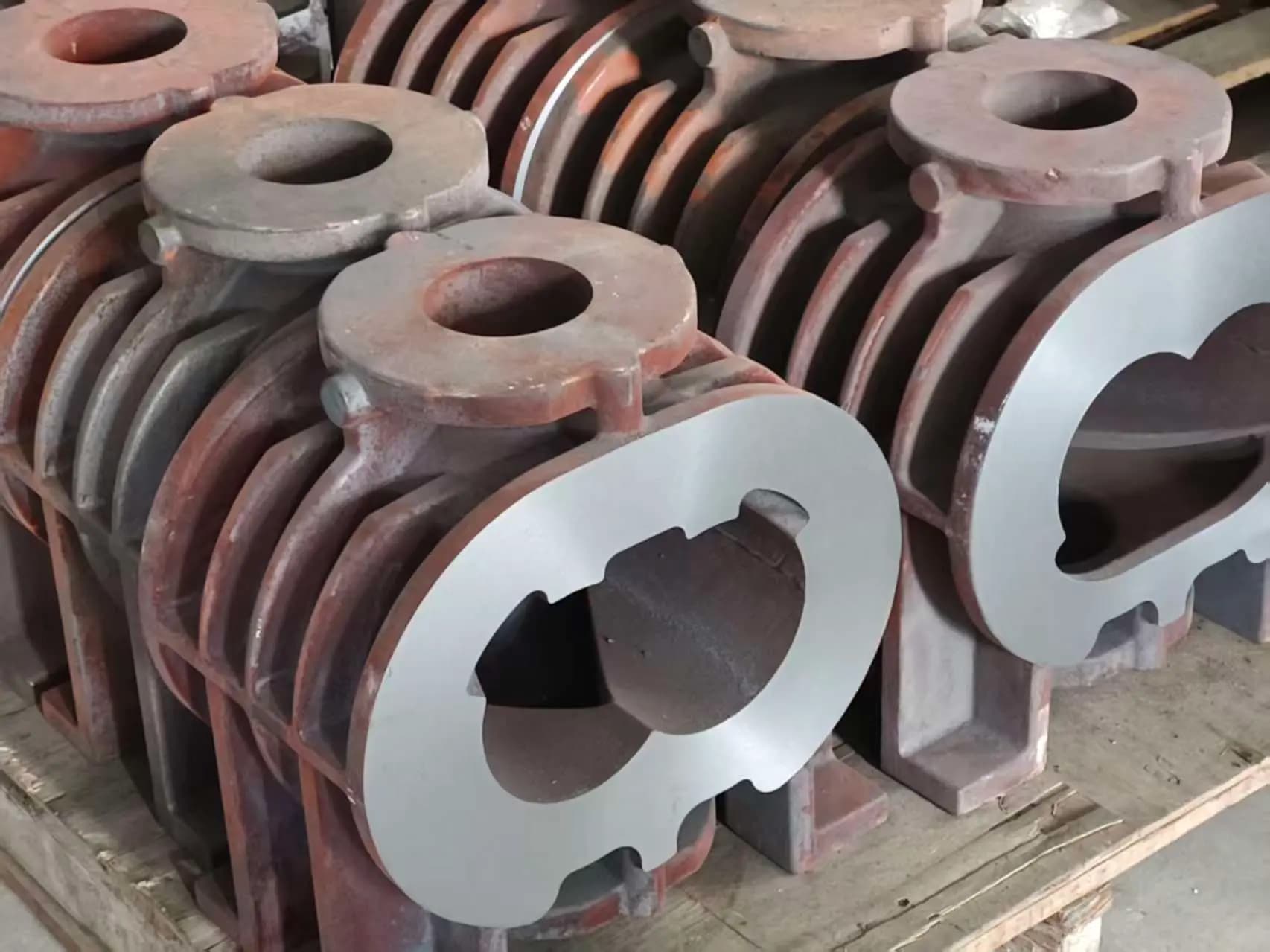 Cast iron dry screw vacuum pump housing blanks staged on the shop floor, one with its sealing face already machined to reveal the twin-bore profile